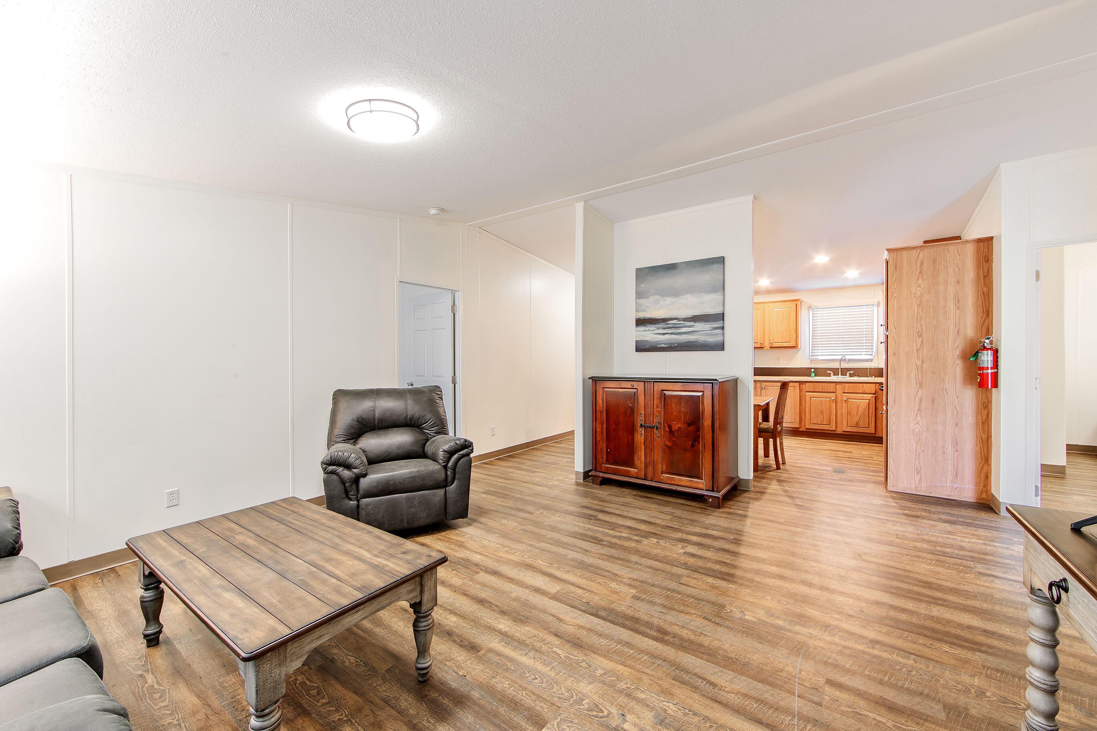 A spacious living room with hardwood flooring, a leather armchair, a wooden coffee table, and a cabinet. A kitchen is visible in the background with wooden cabinets and a window.