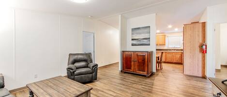 A spacious living room with hardwood flooring, a leather armchair, a wooden coffee table, and a cabinet. A kitchen is visible in the background with wooden cabinets and a window.