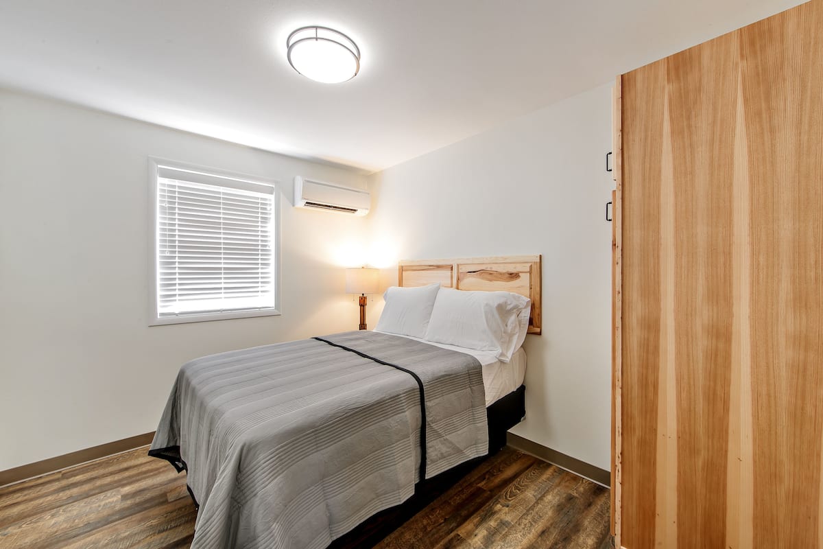 A bedroom with a bed, a bedside table, a lamp, a window with blinds, an air conditioning unit, and a wooden wardrobe.