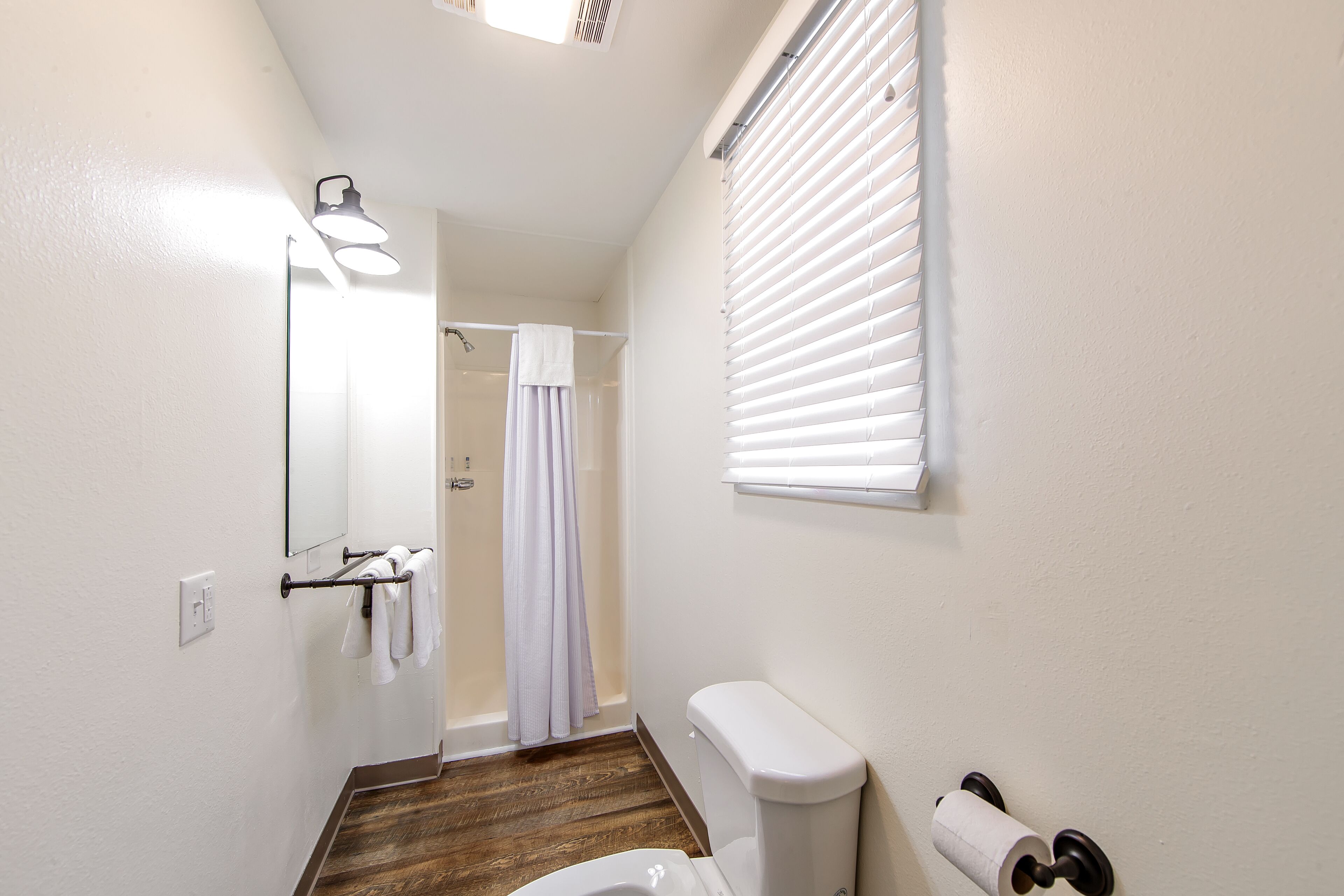 A bathroom with a wooden floor, a white toilet, a towel rack with white towels, a mirror, and a window with blinds.
