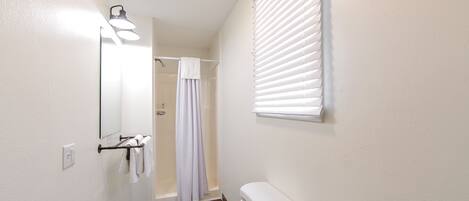A bathroom with a wooden floor, a white toilet, a towel rack with white towels, a mirror, and a window with blinds.
