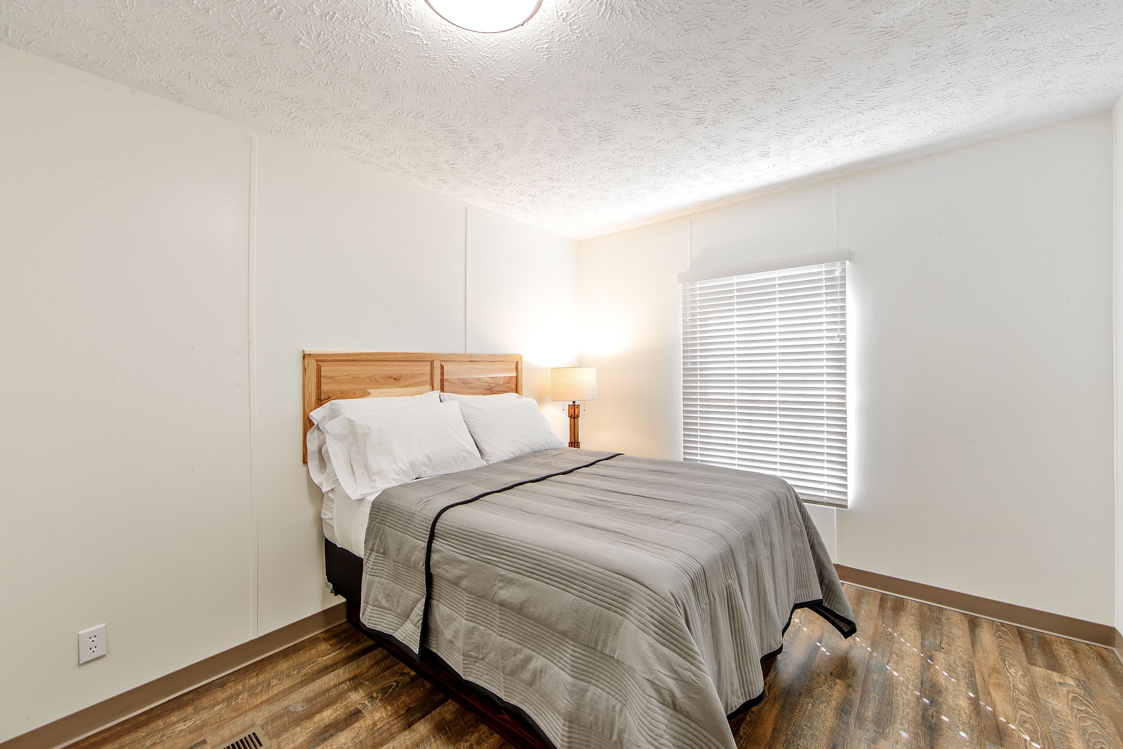 A bedroom with a wooden headboard, a bed with a grey bedspread, a bedside lamp, and a window with blinds.