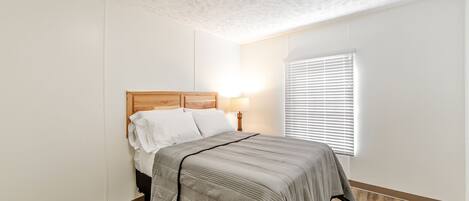A bedroom with a wooden headboard, a bed with a grey bedspread, a bedside lamp, and a window with blinds.