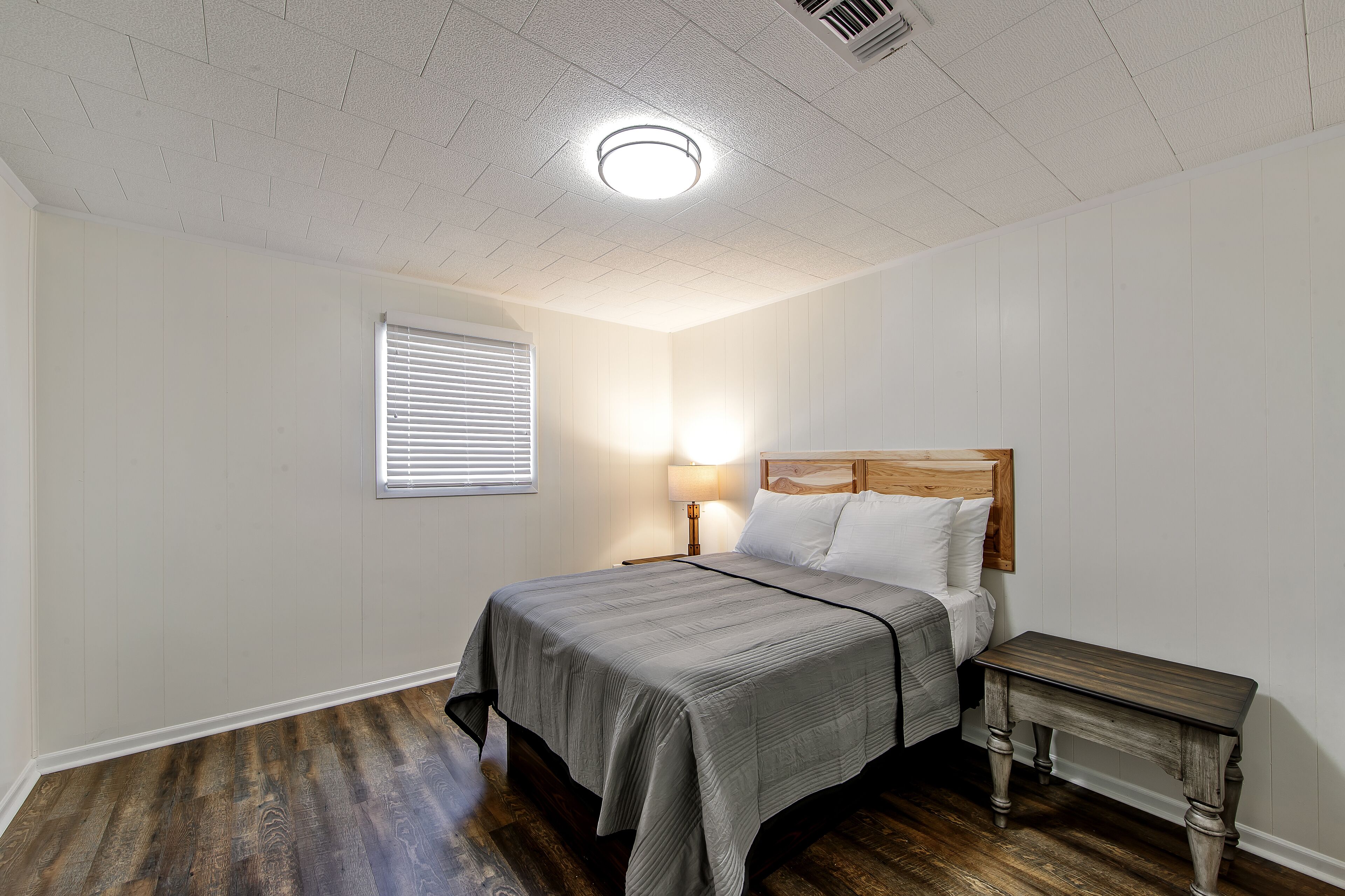 Ein Schlafzimmer mit einem hölzernen Kopfteil, einem Bett mit grauer Bettdecke, einem Nachttisch mit Lampe und einem Fenster mit Jalousien.