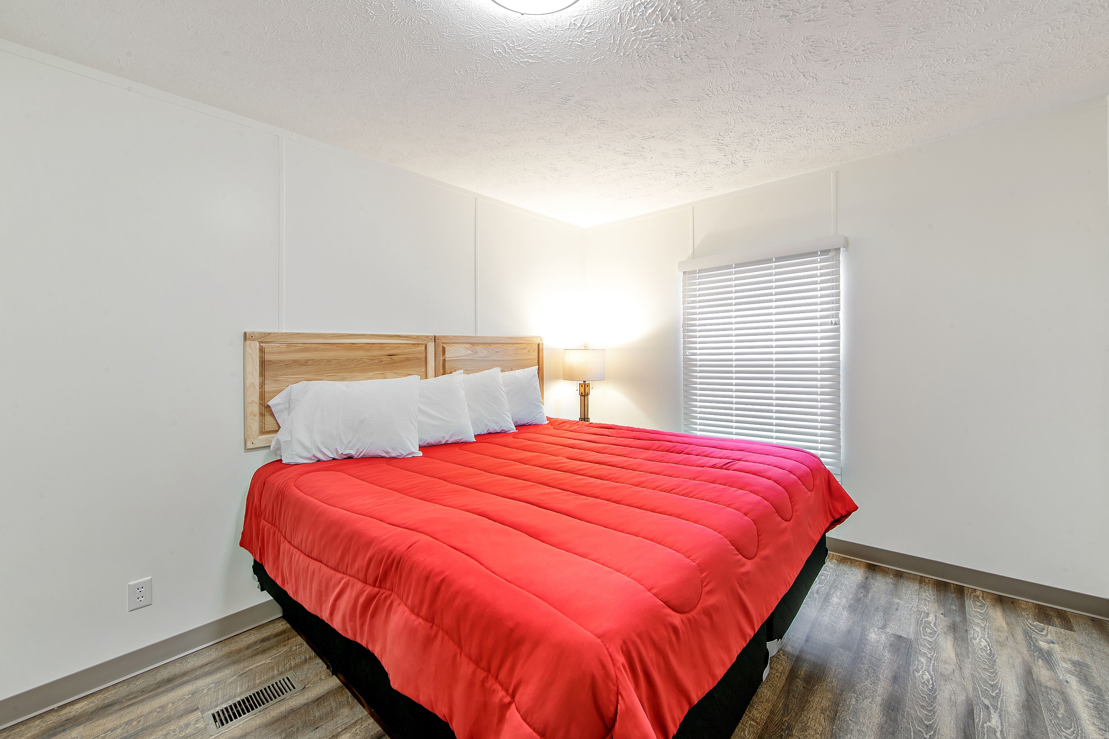 A neatly made bed with a red bedspread and white pillows in a room with white walls and wooden flooring.