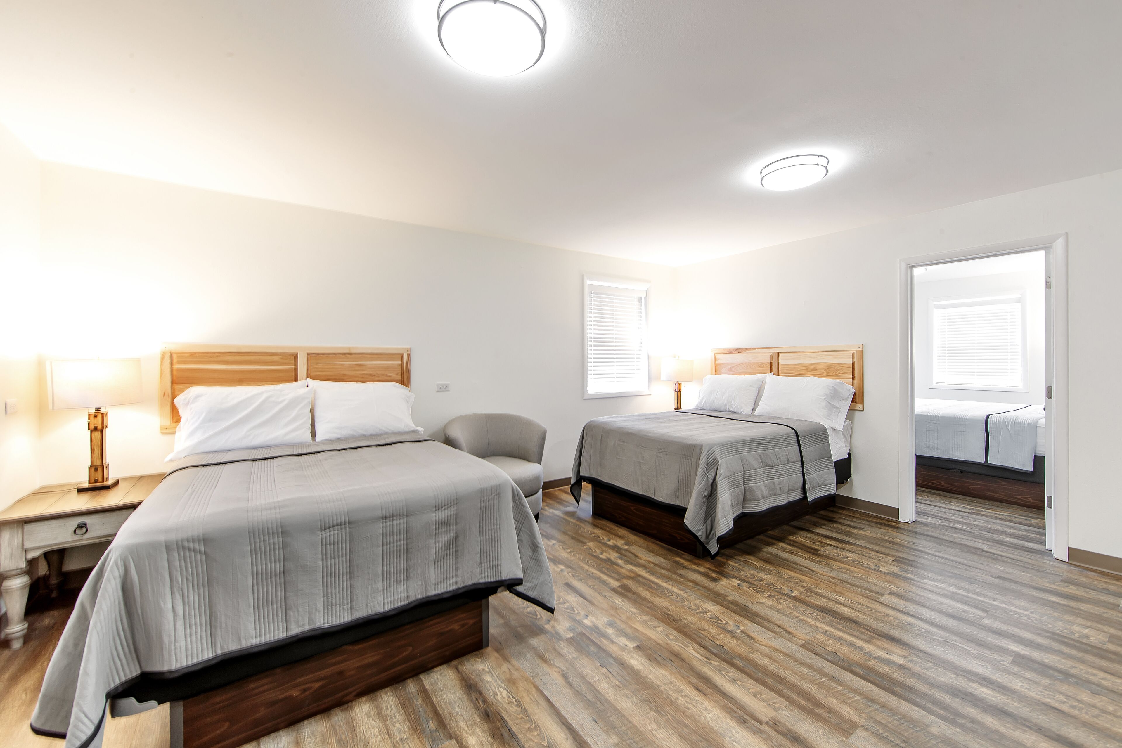 A hotel room with two beds, a wooden floor, and a nightstand with a lamp.