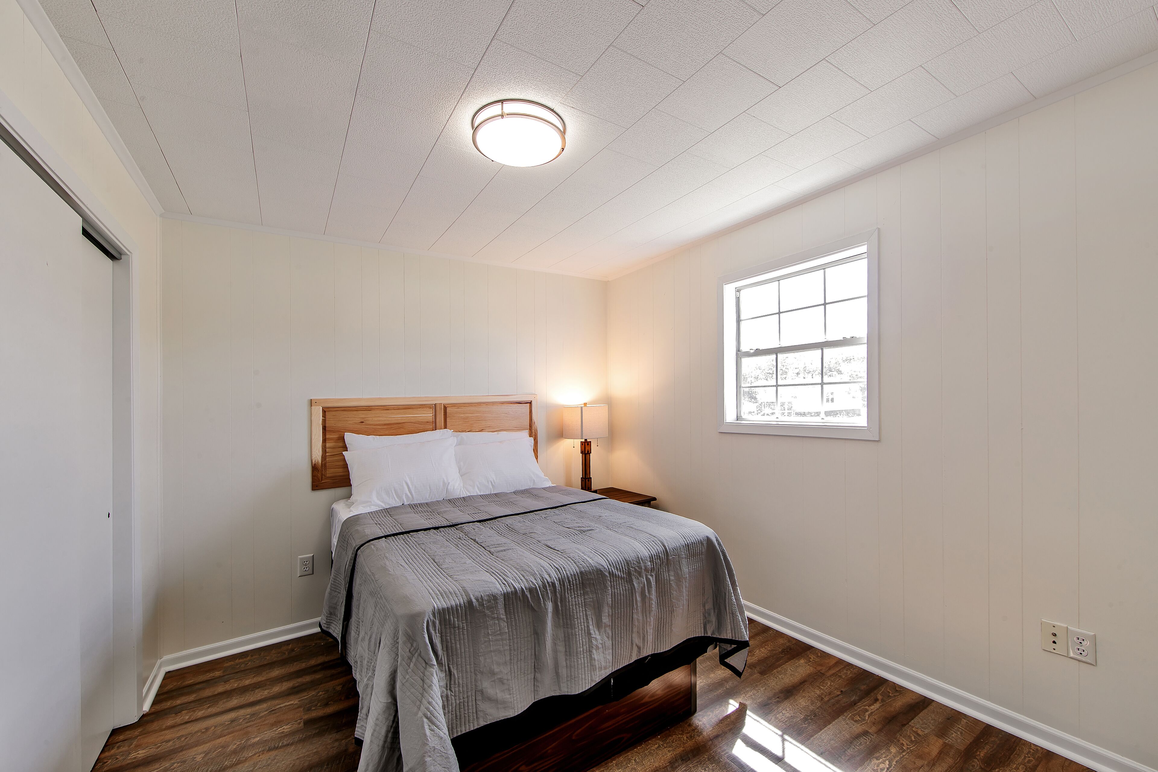 A bedroom with a wooden headboard, a bed with a grey bedspread, a nightstand with a lamp, a window with blinds, and a white ceiling.