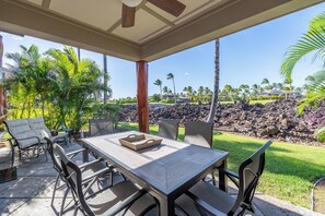 Une terrasse extérieure avec une table et des chaises, un ventilateur de plafond, et une vue sur un espace paysager agrémenté de palmiers et d’une maison en arrière-plan.