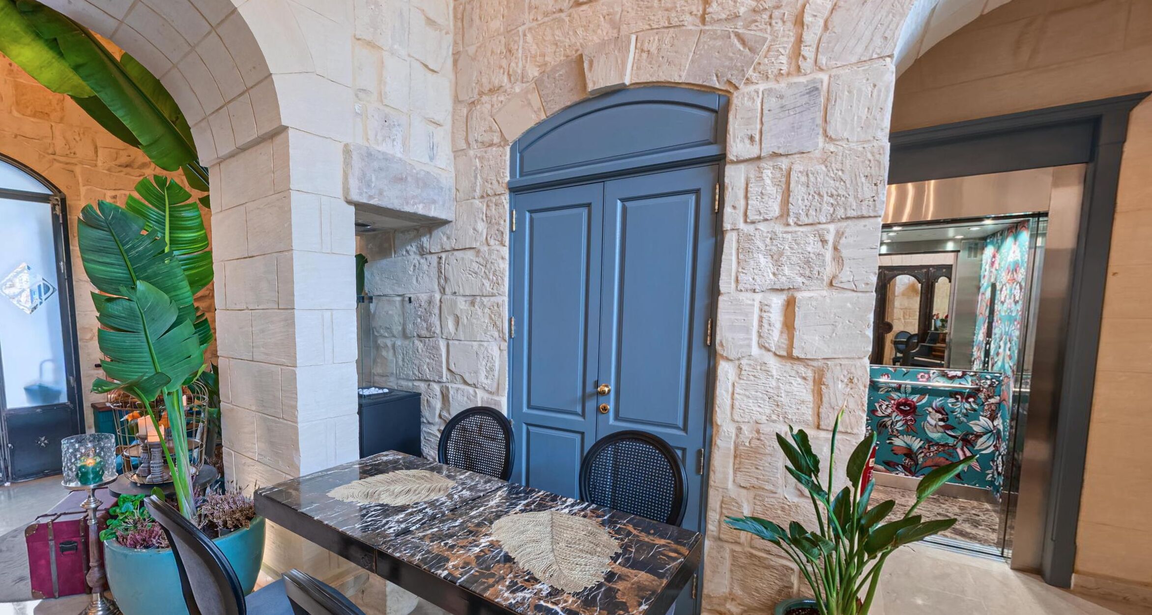 Una habitación con paredes de piedra, una puerta azul, una mesa de mármol y plantas en macetas.