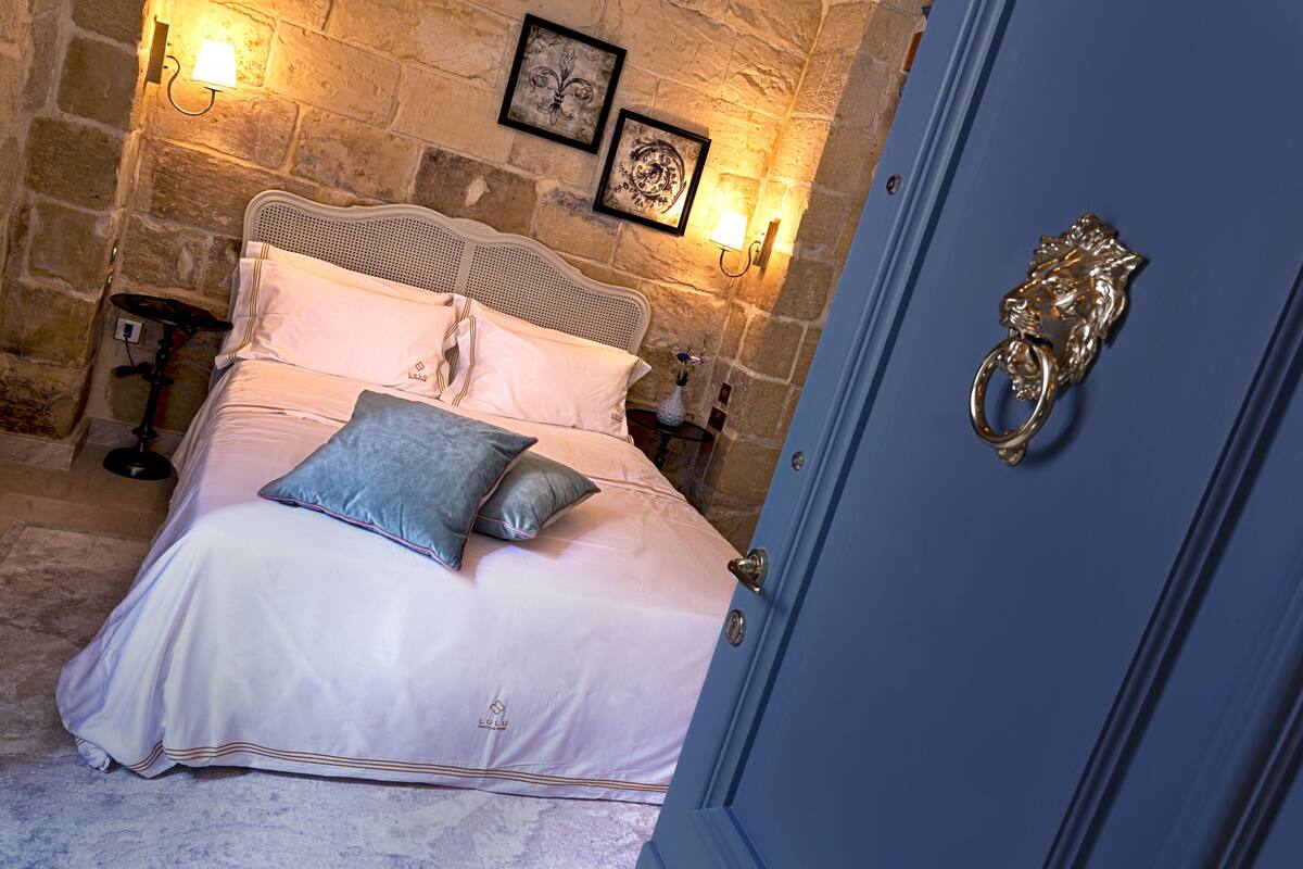 A bedroom with a stone wall, a bed with white bedding, a blue door, and a small table with a lamp.