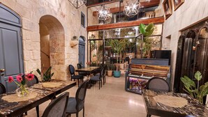 Un restaurante acogedor con paredes de piedra, vigas de madera y un piano. Las mesas están dispuestas con sillas y plantas en macetas.