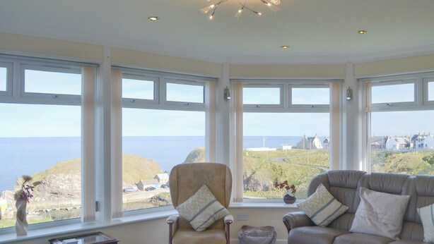 A living room with a large bay window overlooking the sea, a sofa, a chair, and a small table. - Seabreezes - UK30690 (Portknockie)