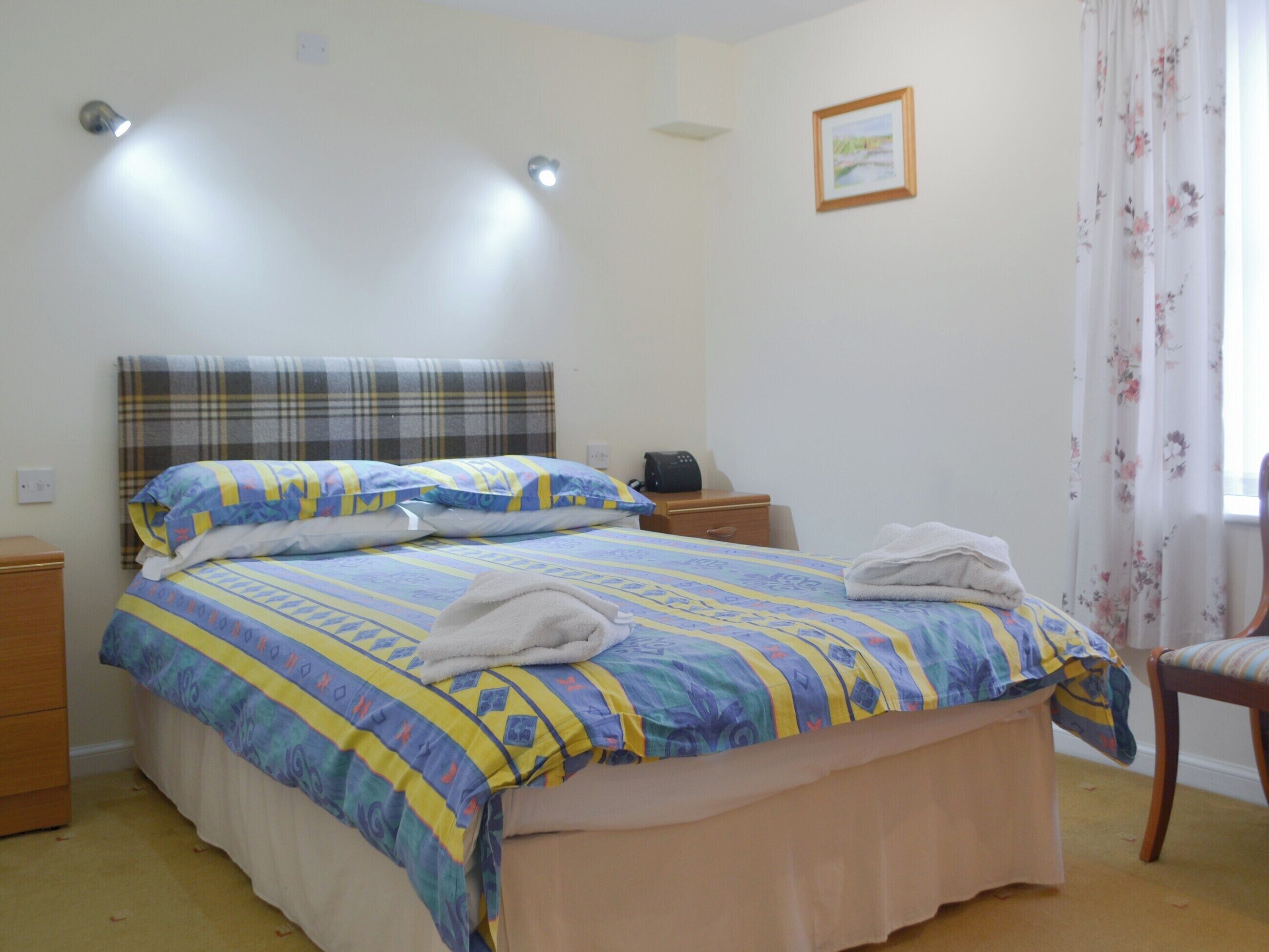 Una cama bien hecha con cabecera de cuadros, una mesita de noche con un teléfono y una silla con cortina estampada.