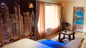 Quarto de hotel com um mural da paisagem urbana, uma cama, uma mesa e vista do oceano através de uma porta.