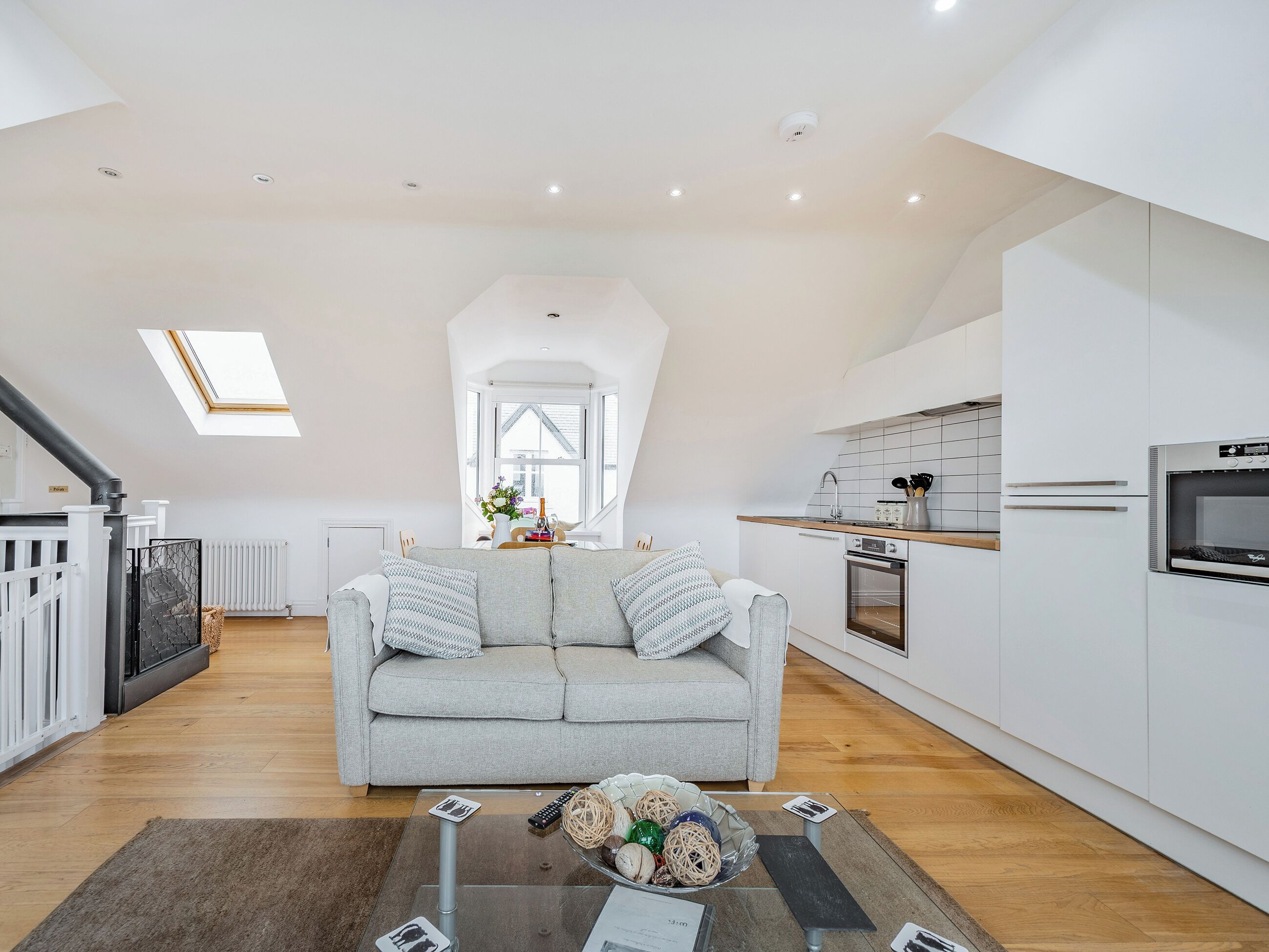 A modern loft apartment with a high ceiling, a grey sofa, a kitchen with white cabinetry, and a skylight.