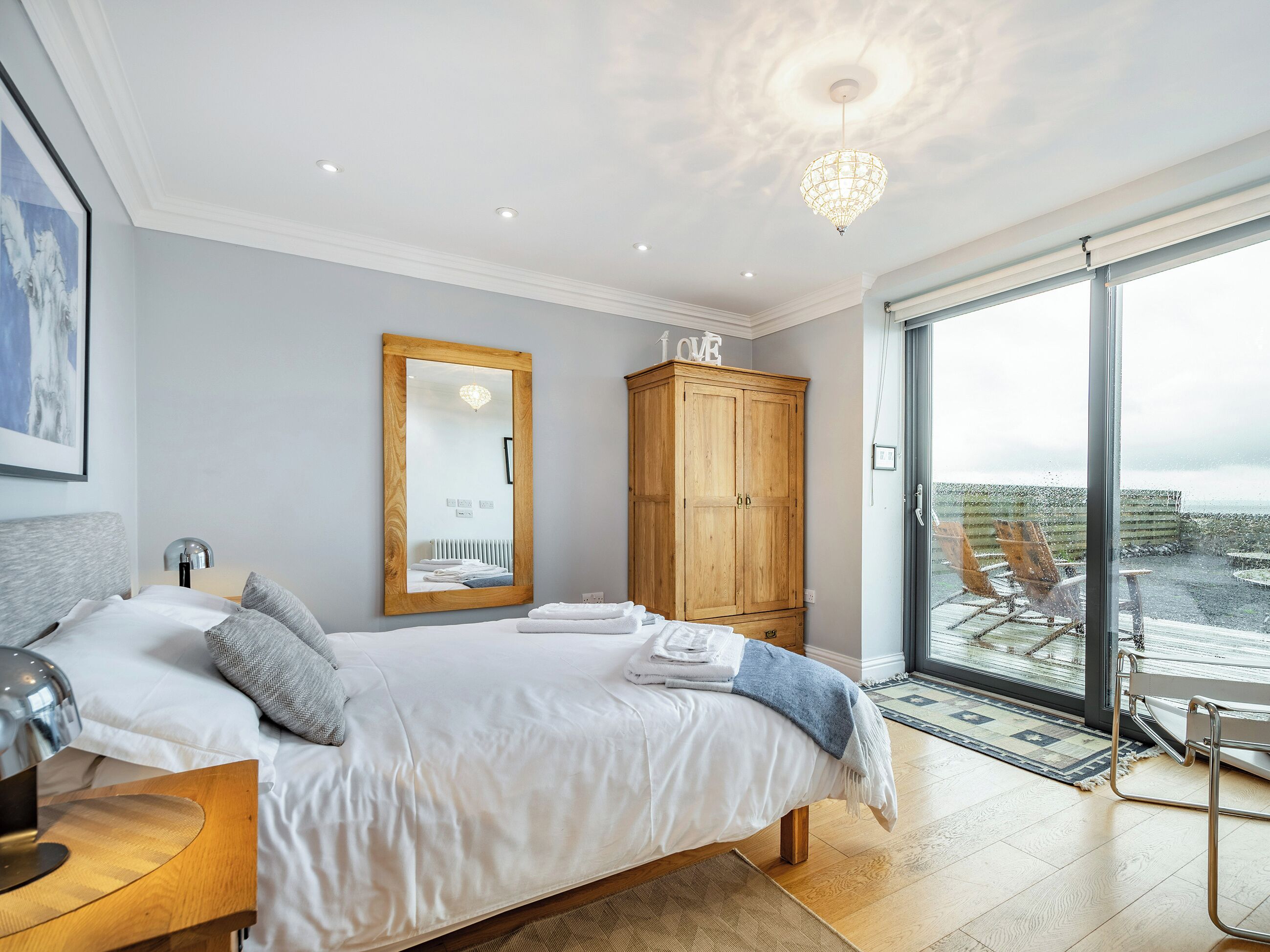 Une chambre à coucher avec un lit, une armoire en bois, un miroir, une lampe de chevet et une vue sur l’extérieur, visible à travers une porte vitrée.