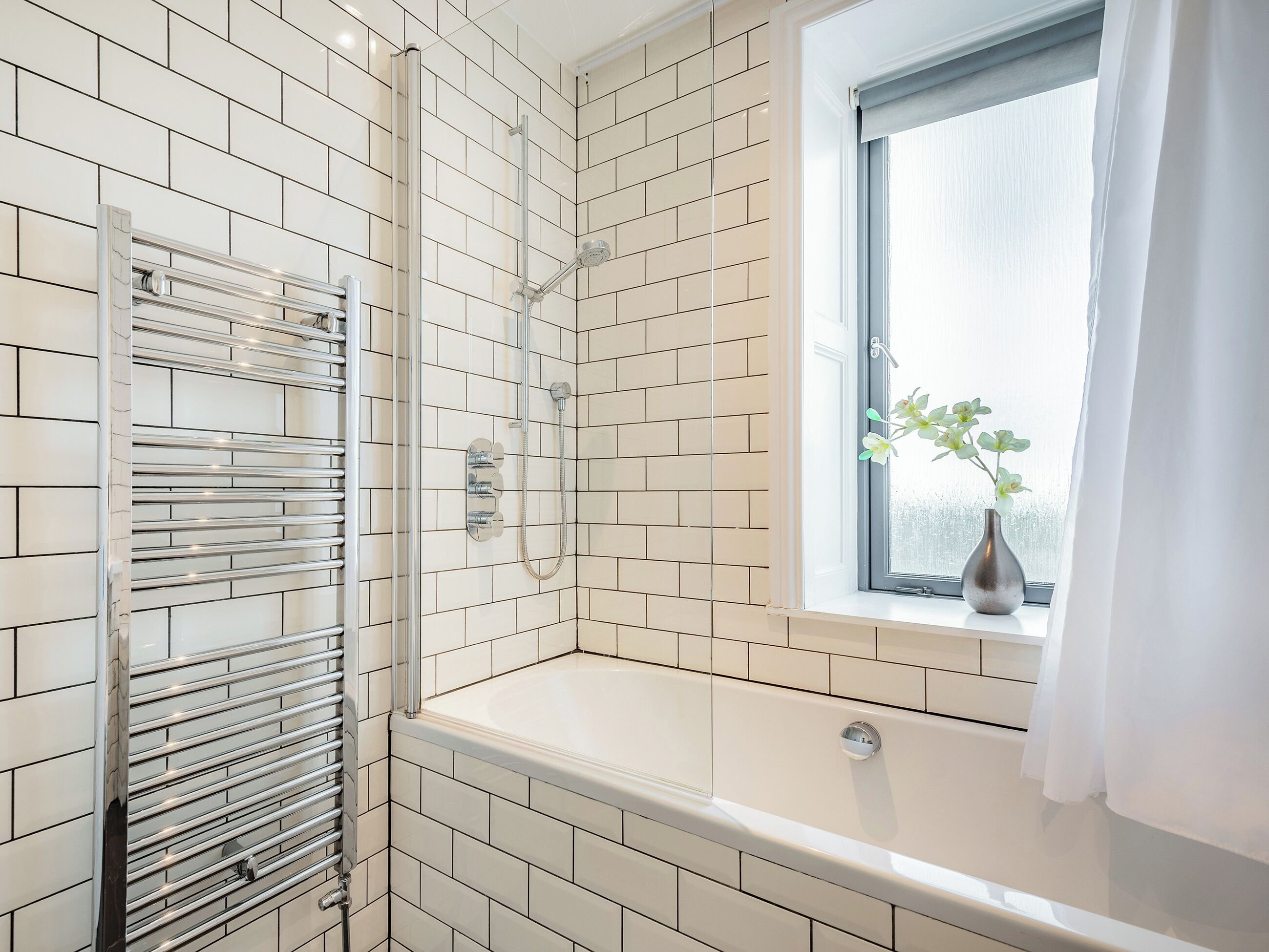A bathroom with white tiled walls, a shower with a glass door, a window with a plant, and a towel rack.