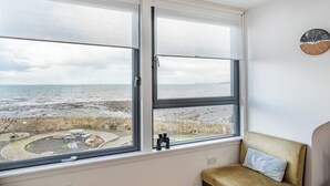 Ett rum med ett stort fönster med utsikt över en strand, en klocka på väggen och en vadderad bänk.