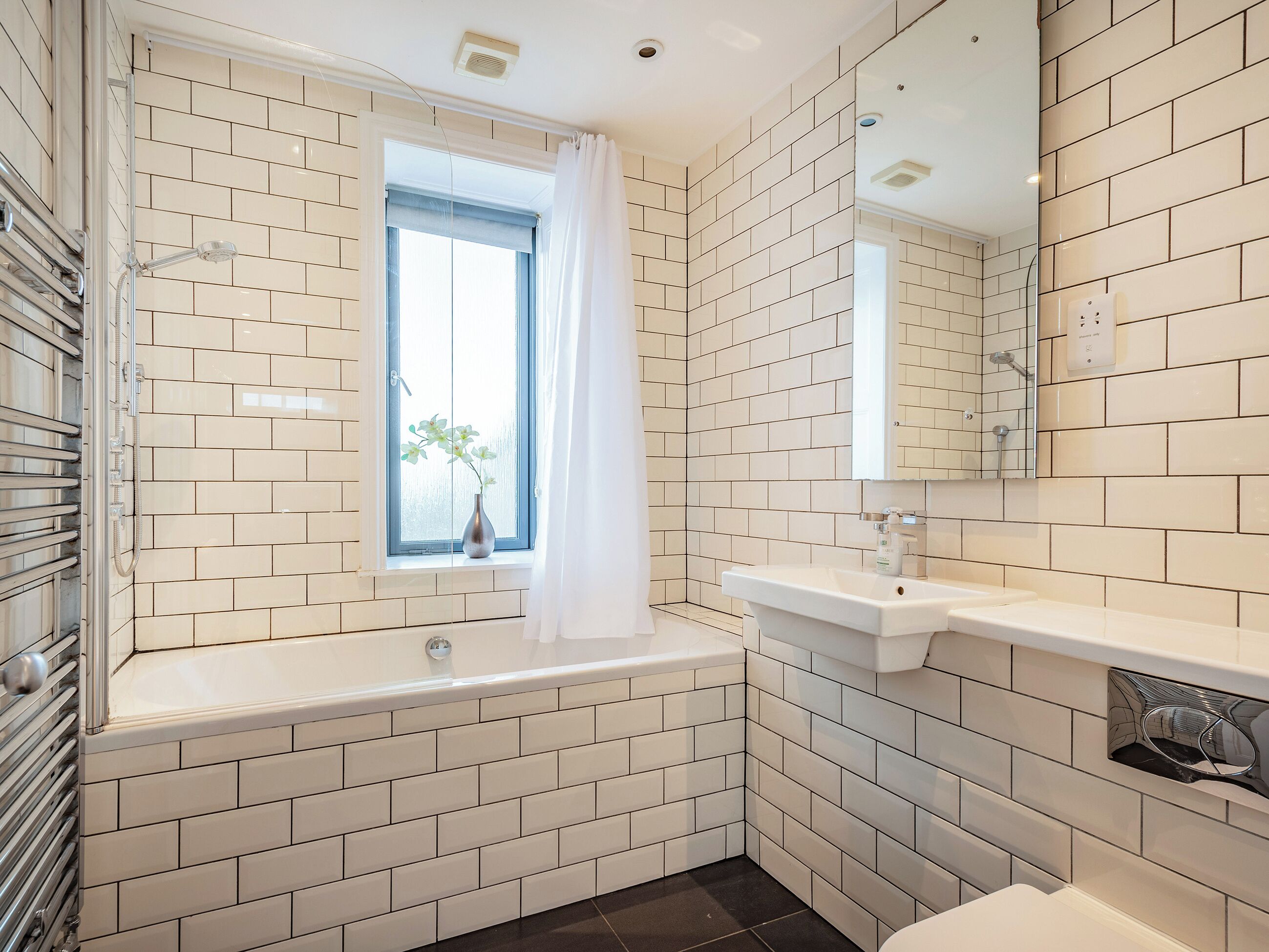 Un baño con azulejos blancos estilo metro, una bañera, un ventanal con cortina, un lavabo y un espejo.