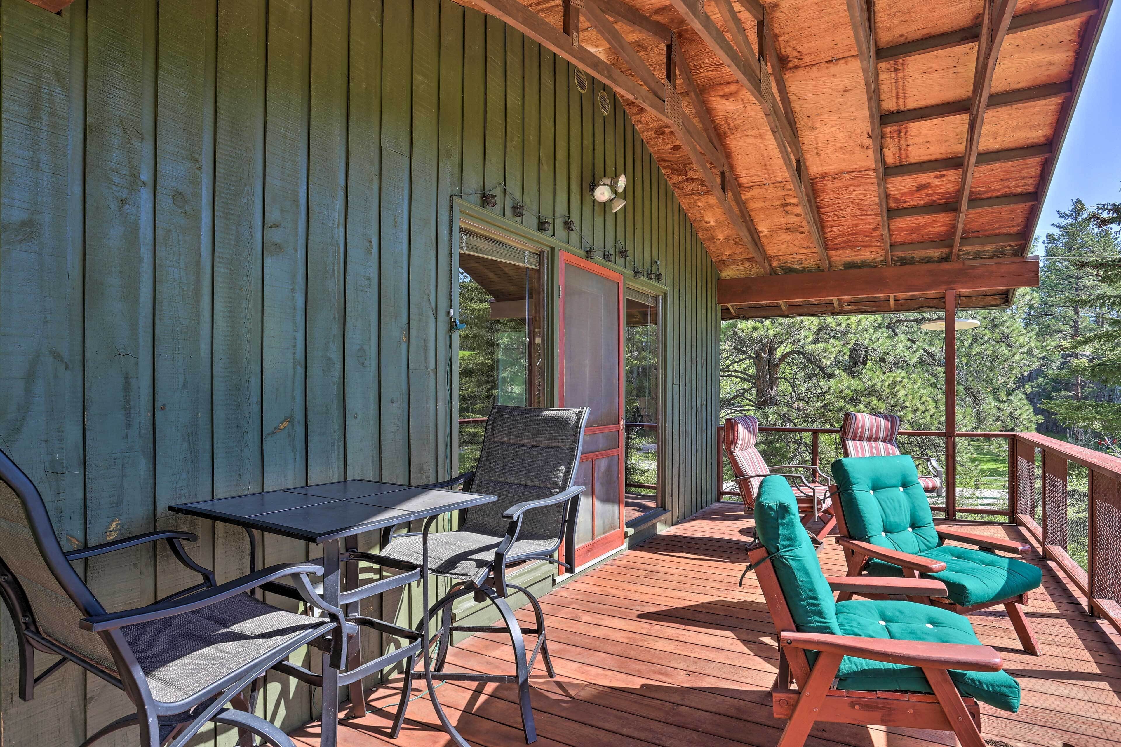A covered outdoor deck with a table and chairs, a wooden railing, and a view of trees.