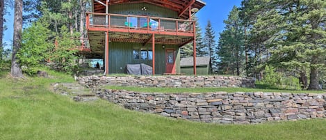 A two-story wooden cabin with a balcony, surrounded by a stone retaining wall and lush greenery.