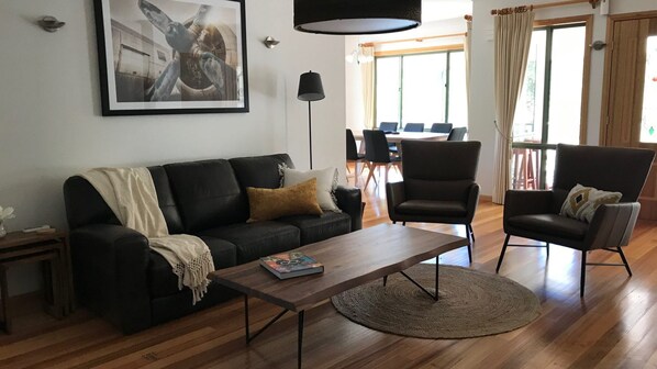 Ein Wohnzimmer mit einer schwarzen Couch, einem Couchtisch und zwei Sesseln. An der Wand hängt ein Gemälde, und im Hintergrund befindet sich ein Essbereich.