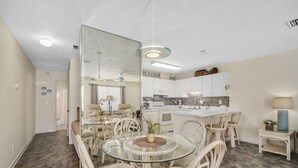 Uma sala de estar ampla com uma mesa de jantar de vidro cercada por cadeiras de vime, uma cozinha com armários brancos e um espelho grande.