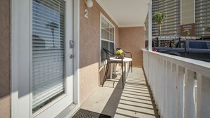 Uma varanda com corrimão branco, uma porta com a placa de número '2', uma pequena mesa com cadeiras e uma palmeira ao fundo.