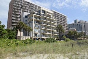 A multi-story hotel with balconies, located near a beach with palm trees and sandy areas.