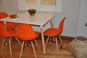 A dining area with a white table, four orange chairs, a bowl of fruit, and a slice of watermelon on the table. - Athens Historic Centre Renovated Apartment (Athina)