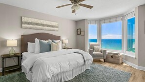 A bedroom with a large bed, a ceiling fan, a view of the ocean, and a chair by the window. - Gulf-Front 2 Bed, 2 Bath • Sleeps 8 • Amenity-Packed • Beach Chair Service for 2 (Panama City Beach)