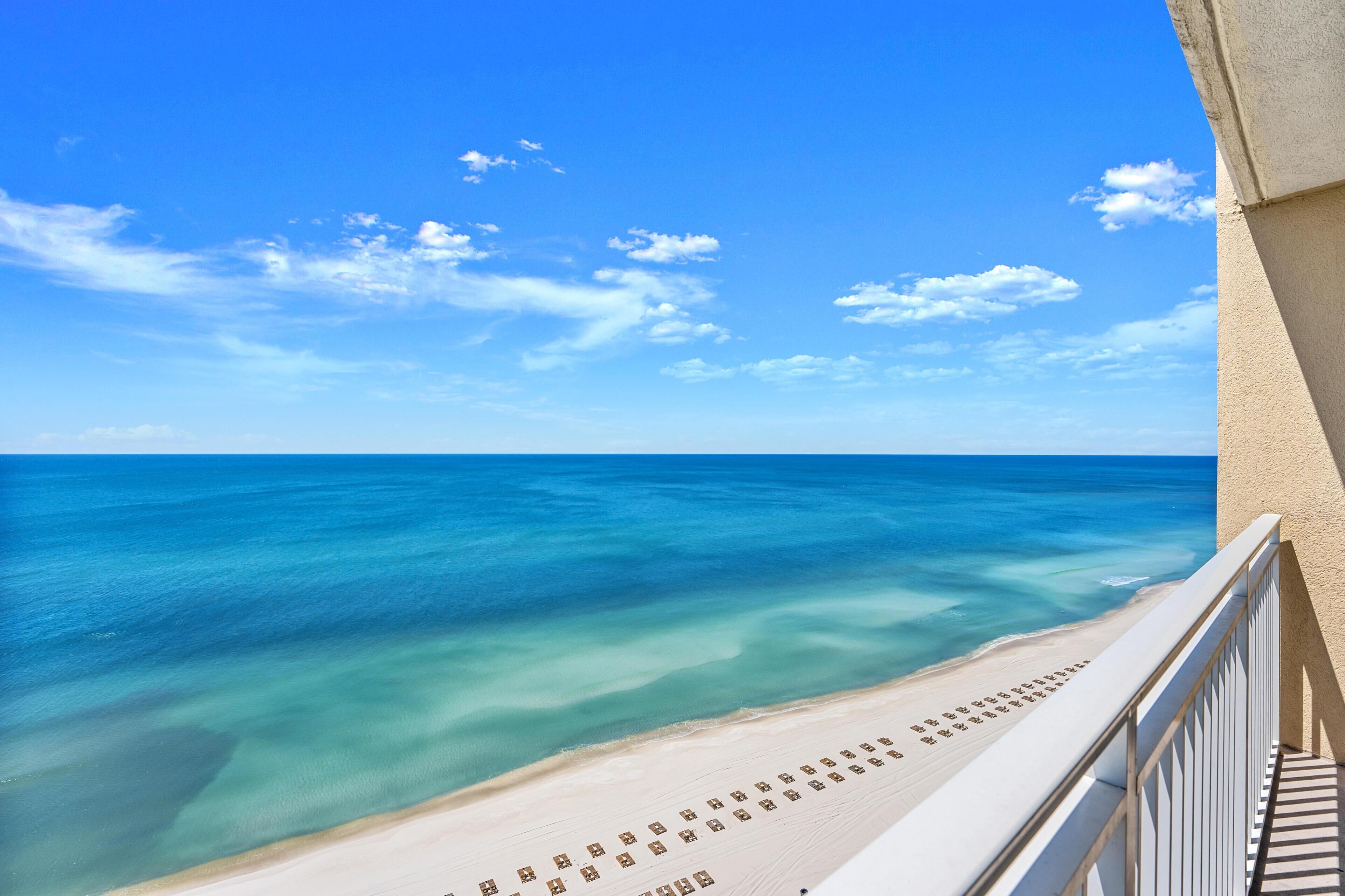 Blick auf den Strand von einem Balkon mit Geländer, klarer, blauer Himmel und ruhiges Meer.