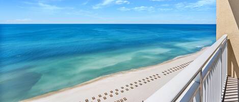 Blick auf den Strand von einem Balkon mit Geländer, klarer, blauer Himmel und ruhiges Meer.