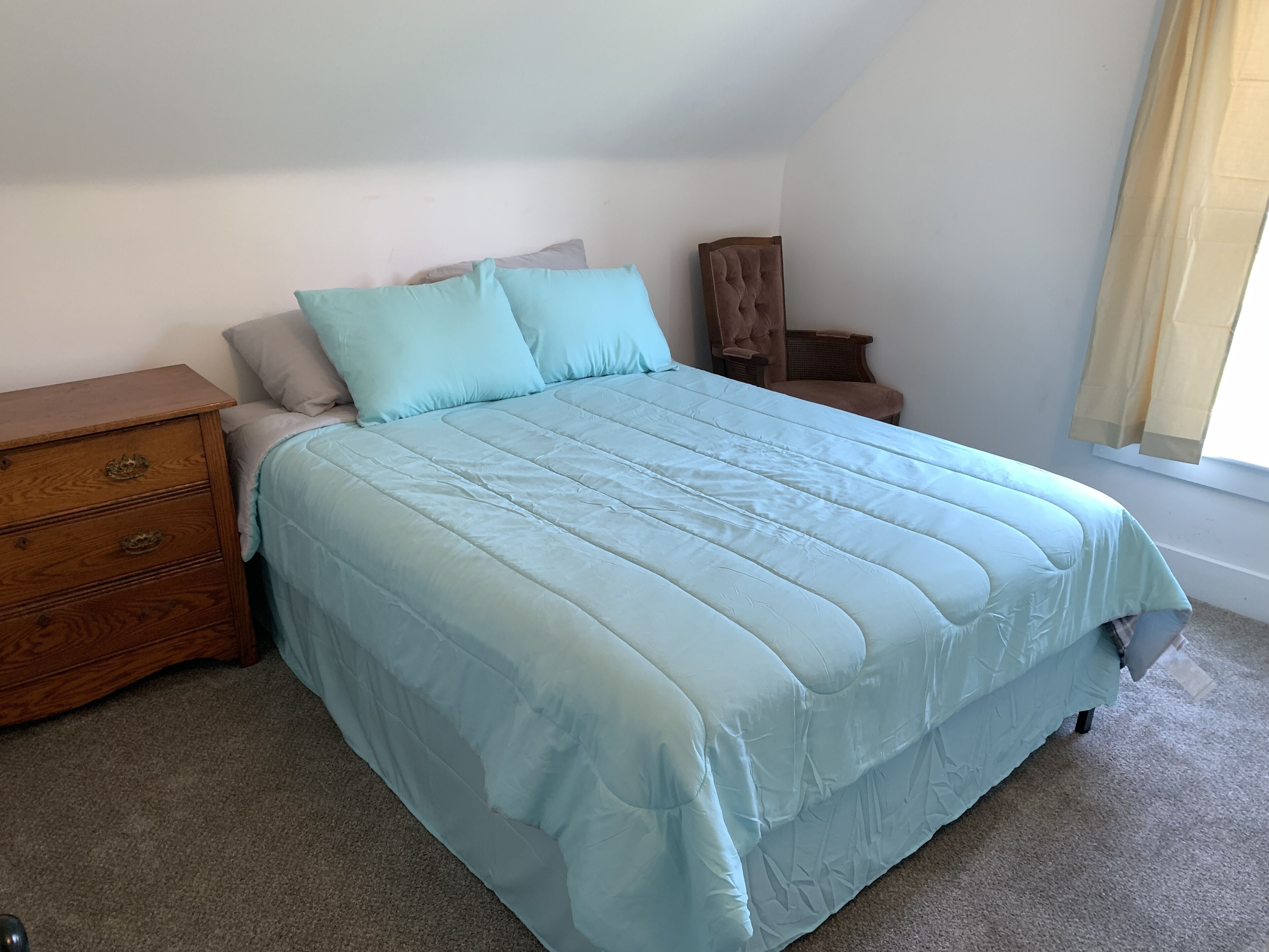 Une chambre à coucher avec un lit, une table de chevet en bois, une chaise et une fenêtre avec des rideaux.