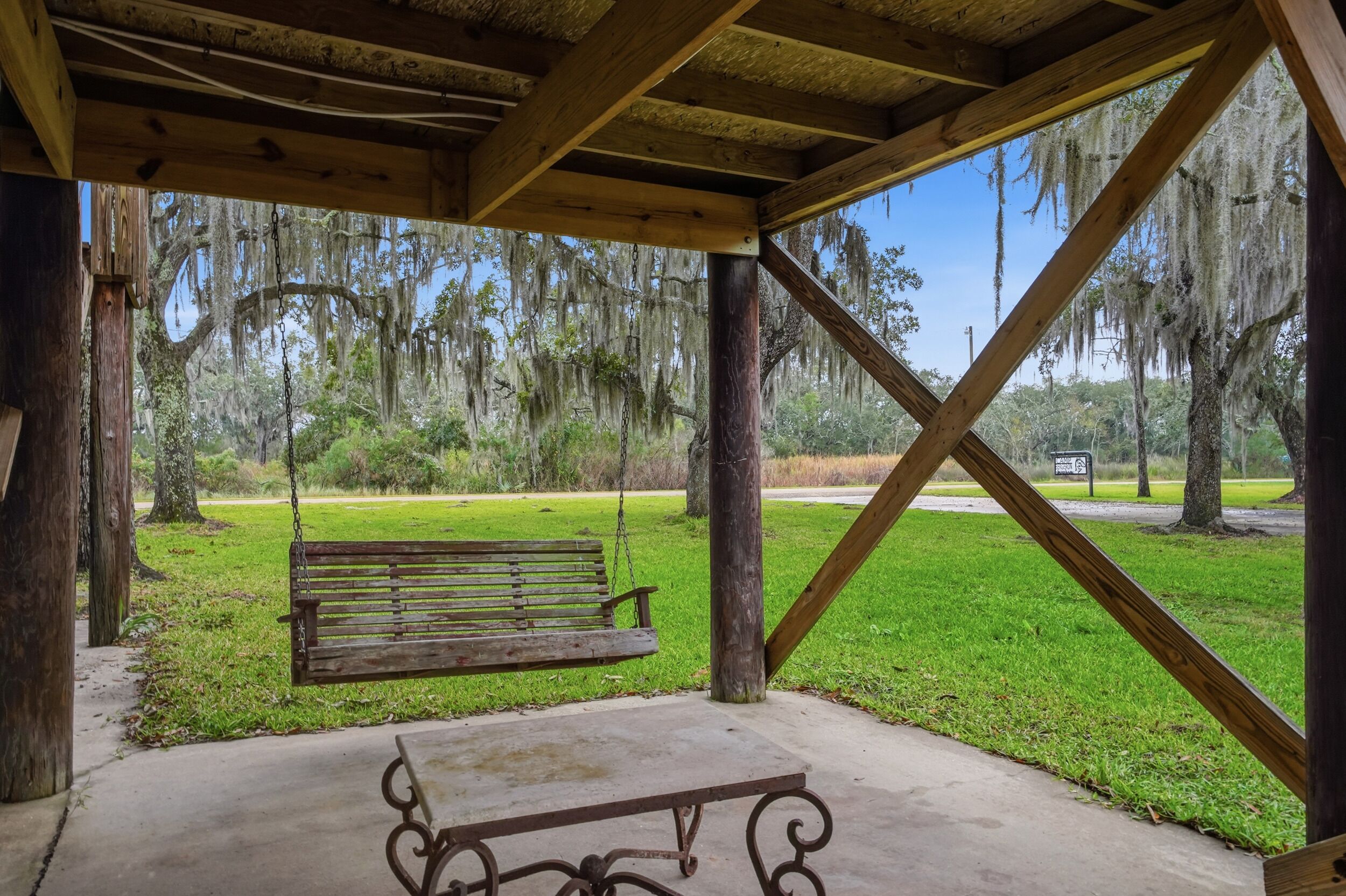 Un quiosco de madera con banca y mesa, con vista a un parque con árboles y cielo despejado.
