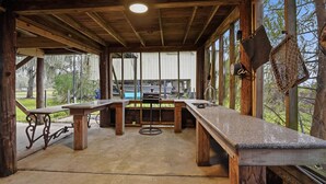 A covered outdoor dining area with wooden tables and benches, a concrete floor, and large windows offering views of the surrounding landscape. - Beautiful, Relaxing, Spacious Cottage with view of Vermillion Bay! (Cypremort Point)