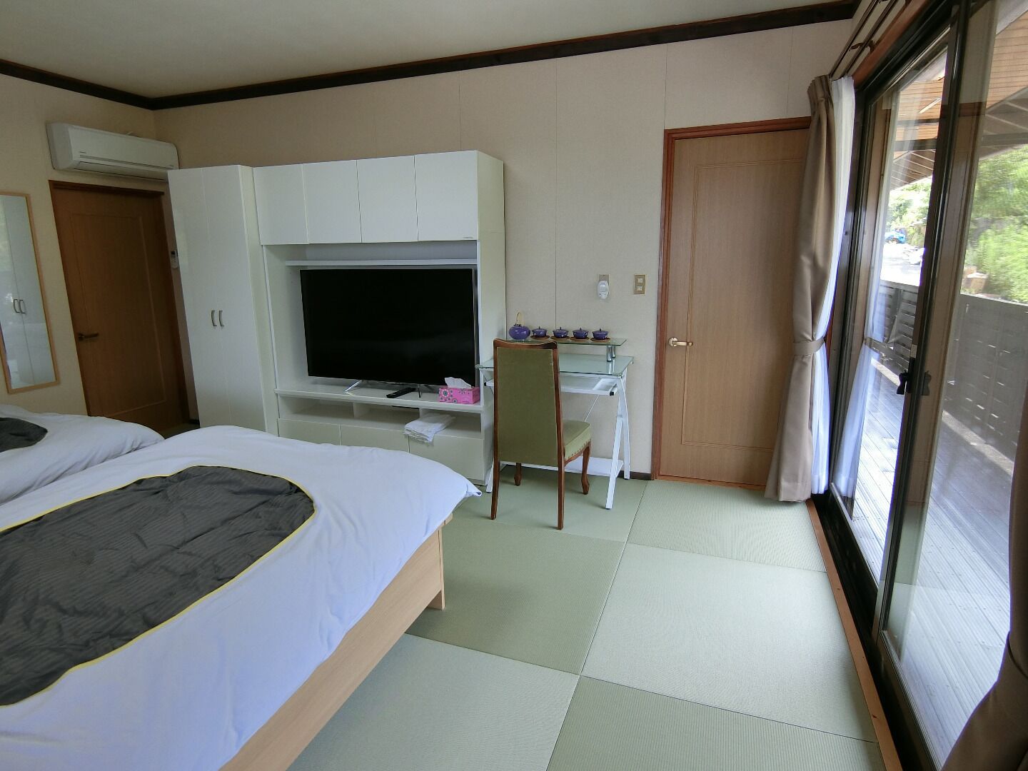 Habitación de hotel con cama, escritorio con televisor, silla y puerta corrediza a un balcón.