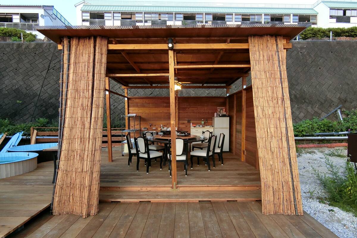 Une terrasse extérieure en bois, dotée d’un espace repas comprenant une table et des chaises, ainsi que d’un coin cuisine avec un réfrigérateur et des placards.