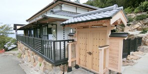 Een traditioneel Japans gebouw met een houten poort, een stenen muur en een balkon met uitzicht op een rotsachtige heuvel.