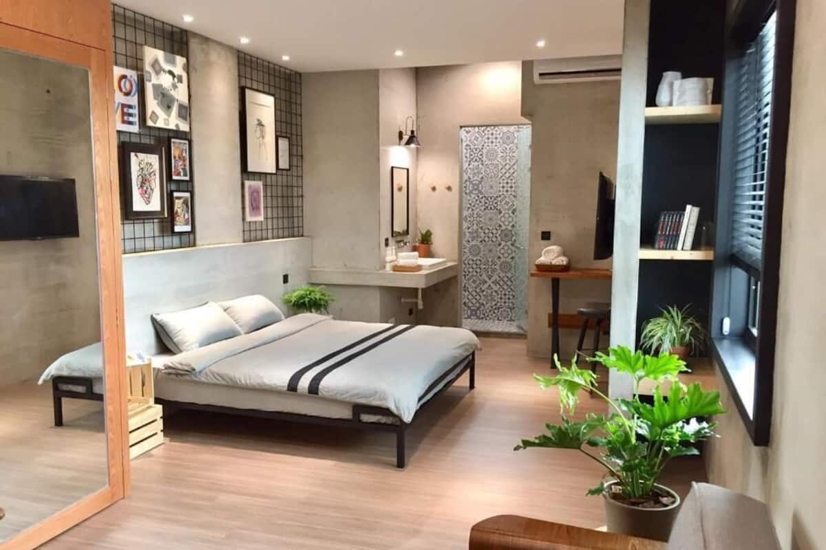 A modern bedroom with a bed, bedside table, and a small desk with a chair. There are potted plants and a wall-mounted TV.