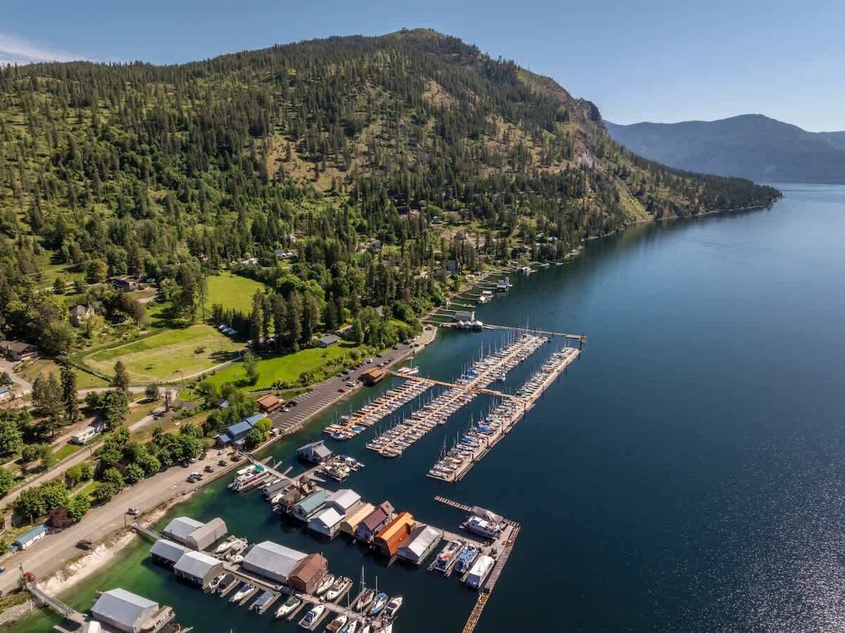 Une vue aérienne d’une marina où sont amarrés des bateaux, entourée de collines boisées et surplombée par un ciel dégagé.