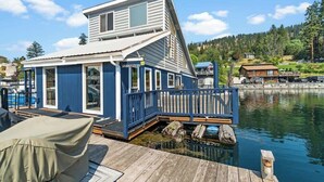 A houseboat with a deck and a covered seating area, docked at a calm body of water. - Lakefront Float Home with Huge Deck & Water Toys (Bayview)