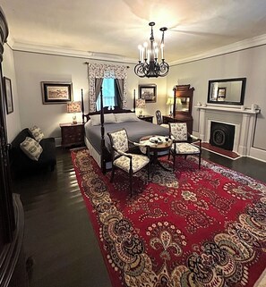 A bedroom with a four-poster bed, a chandelier, a fireplace, and a red patterned rug.