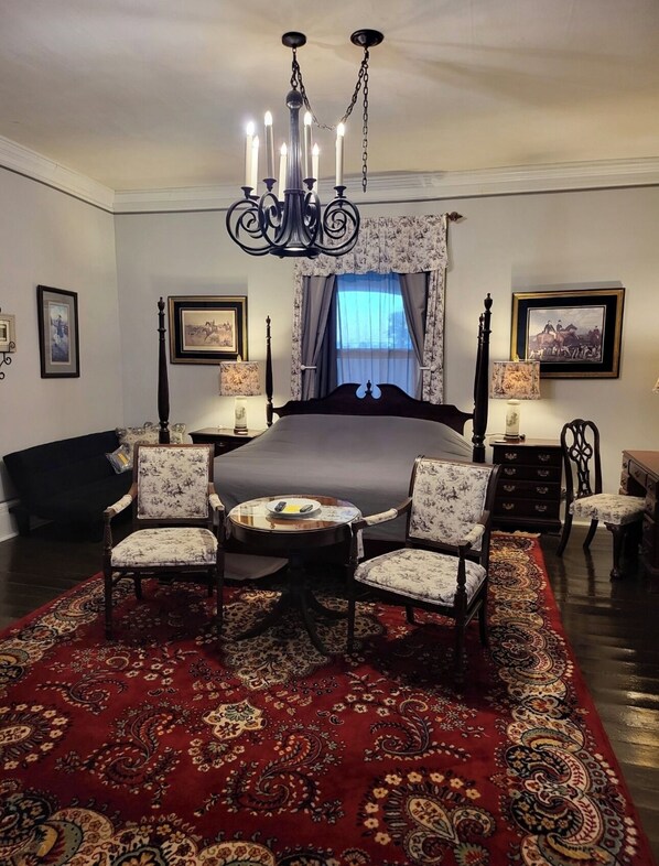 A bedroom with a large bed, two chairs, a small table, a chandelier, and a red patterned rug.