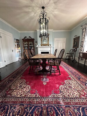 A dining room with a large wooden table, ornate chairs, a red patterned rug, and a chandelier.