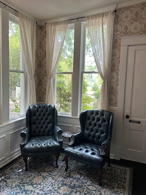 Um quarto com duas poltronas de couro preto, um tapete estampado e vista para o verde através de grandes janelas com cortinas estampadas.