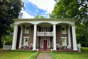 A two-story brick house with a central balcony, white columns, and a front porch with outdoor seating.