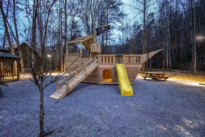 A wooden play structure with a slide and stairs, surrounded by trees and a picnic table.