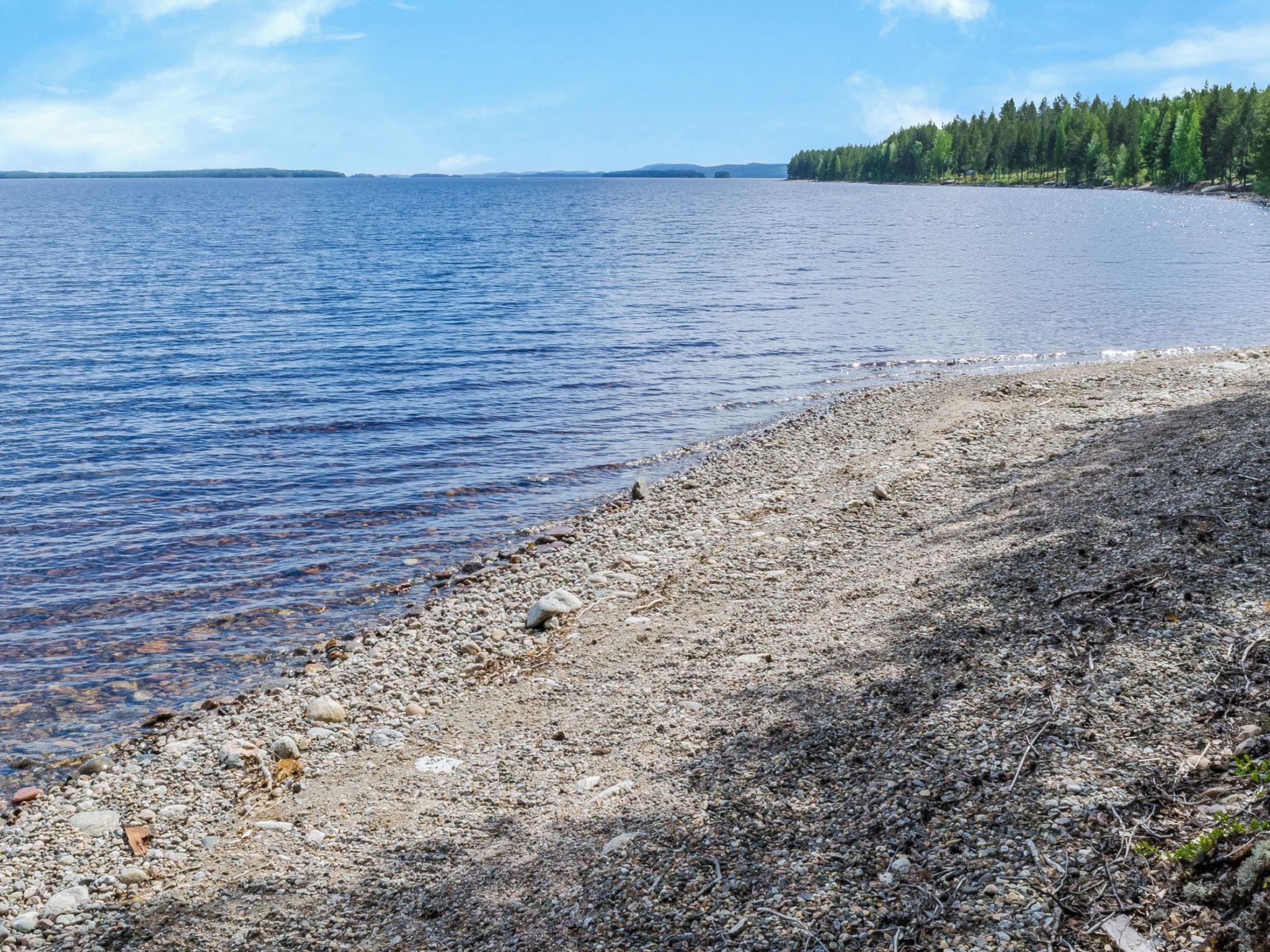 Ein kiesiger Strand mit ruhigem Wasser und einer bewaldeten Insel in der Ferne.