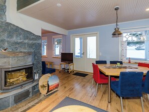 Una sala de estar moderna con chimenea de piedra, un comedor con mesa de madera y sillas azules, y un ventanal con vistas a árboles.